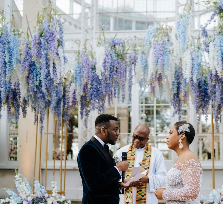 Bride in illusion v-neck sheer off shoulder long sleeve pearl embellished wedding gown standing with groom, reading his vow renewal, wearing velvet midnight blue double breasted groom’s blazer, velvet bowtie and peach garden rose boutonniere underneath suspended pastel flower arrangements at Syon Park