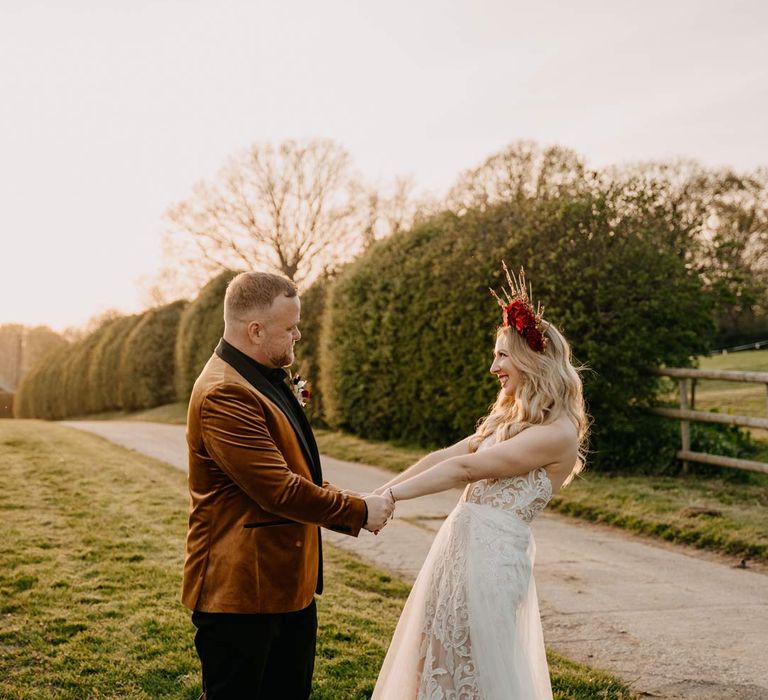 Groom in crushed velvet burnt orange grooms blazer and dried flower boutonniere holding hands with bride in off shoulder lace wedding dress with corset style top and red rose and gold halo bridal crown on the grounds of Southlands Barn wedding venue