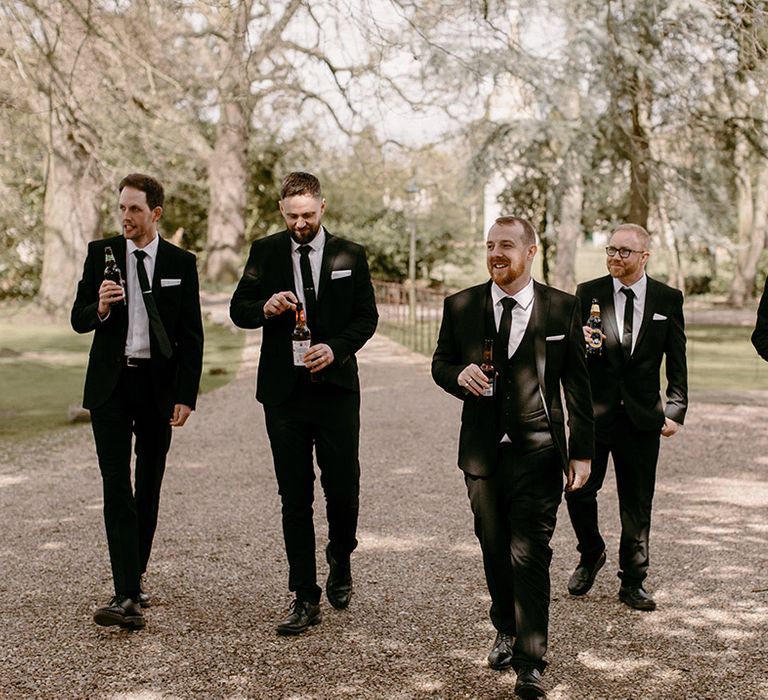 Groomsmen in black and white traditional wedding suits walking together with bottles of beer and drinks 