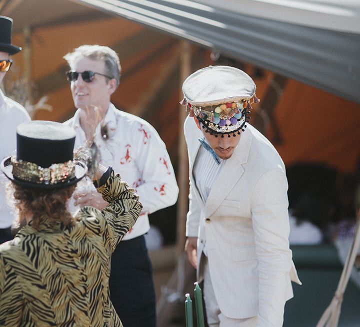 Wedding guest in white suit with white boho hat featuring multicoloured pompoms