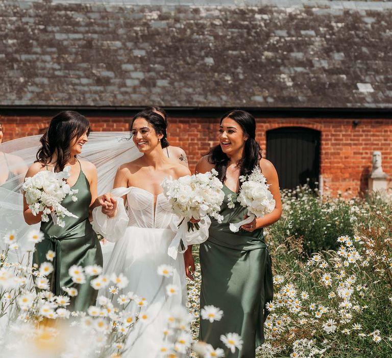 bride in off-the-shoulder tulle wedding dess and bridesmaids in green satin bridesmaid dresses walk through a daisy garden whilst holding white rose bouquets