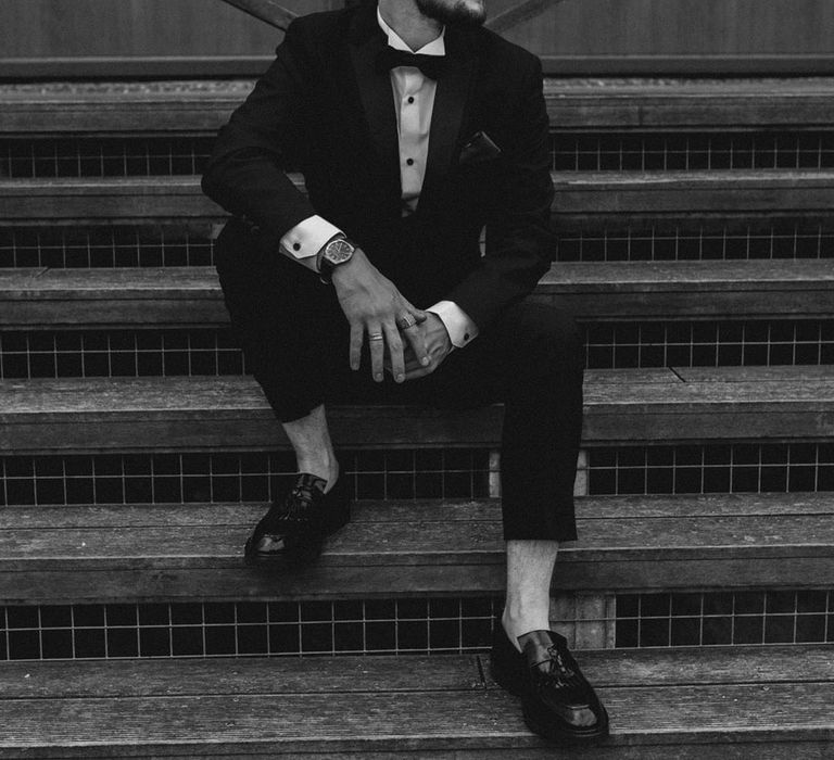 Groom sits on the steps to the glass house wedding venue in his black tie for the traditional wedding 