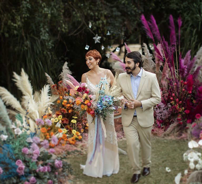 Bride wearing lace wedding dress carrying a multicoloured rainbow wedding bouquet walking back down the aisle with the groom 