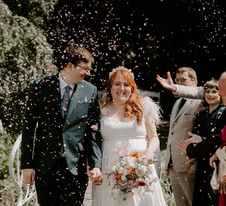 Bride wearing white flower headpiece with feather wedding dress walking with the groom in a black suit with purple tie for confetti moment 