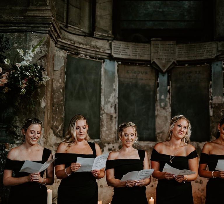 The bridal party wearing black bridesmaid dresses perform a wedding reading together 