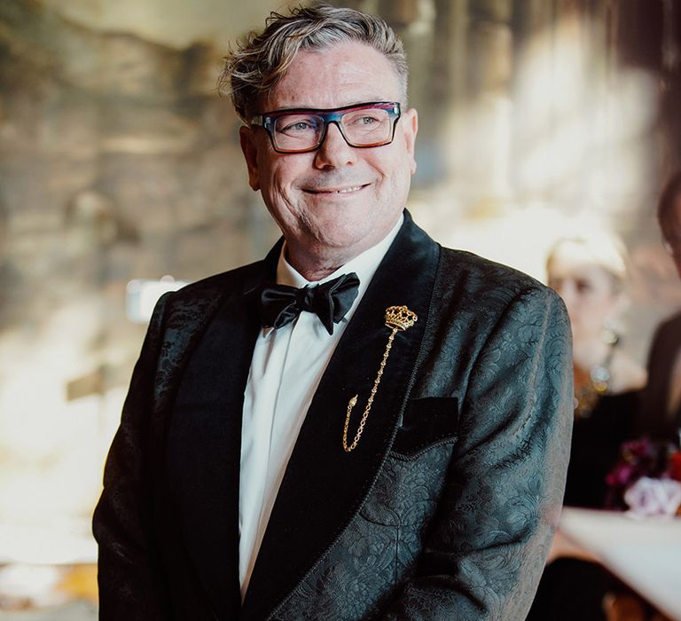 Groom in all black brocade and velvet tuxedo with unique gold crown pin with black glasses standing for the wedding ceremony 