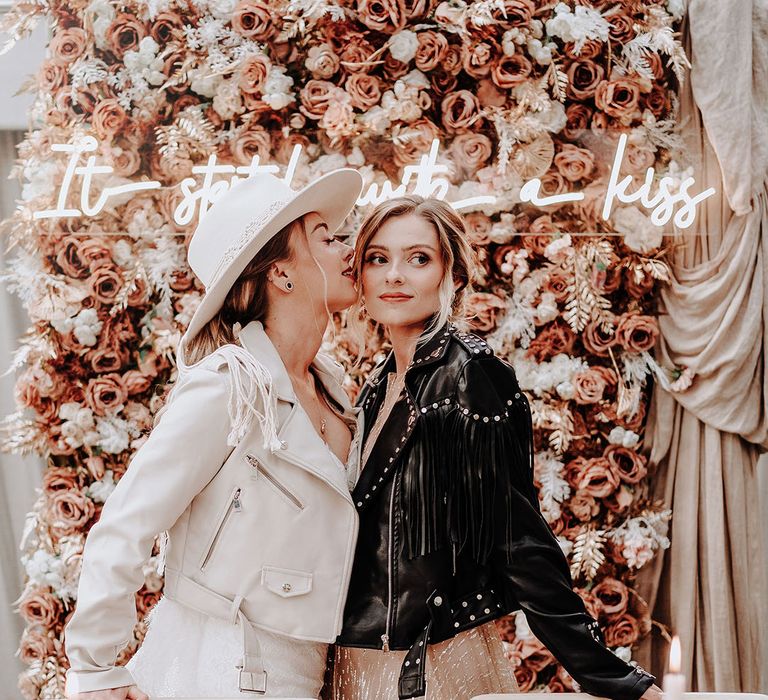 Two brides wearing personalised leather jackets in front of pink flower wall with wedding hat