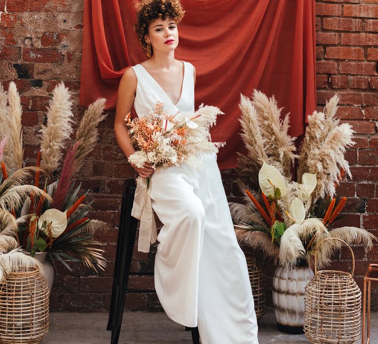 stylish bride in wedding jumpsuit sitting on a stool 