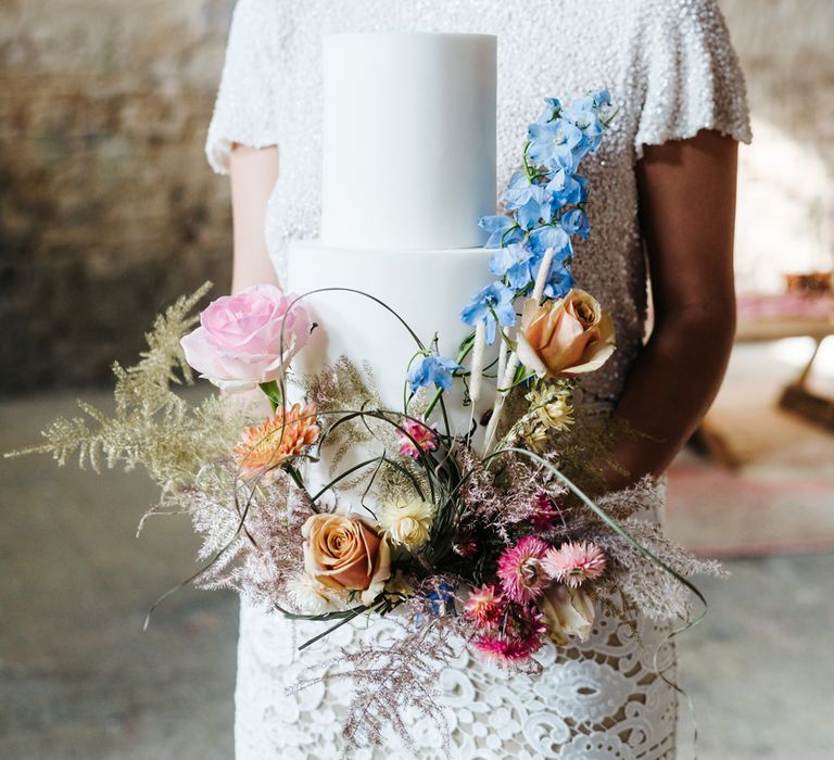 Bride holding a white iced wedding cake decorated with colourful flowers 