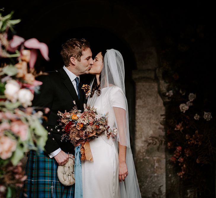 A bride and groom kiss near Christmas church wedding flowers. The groom wears a kilt and the bride wears an embellished white dress and veil.