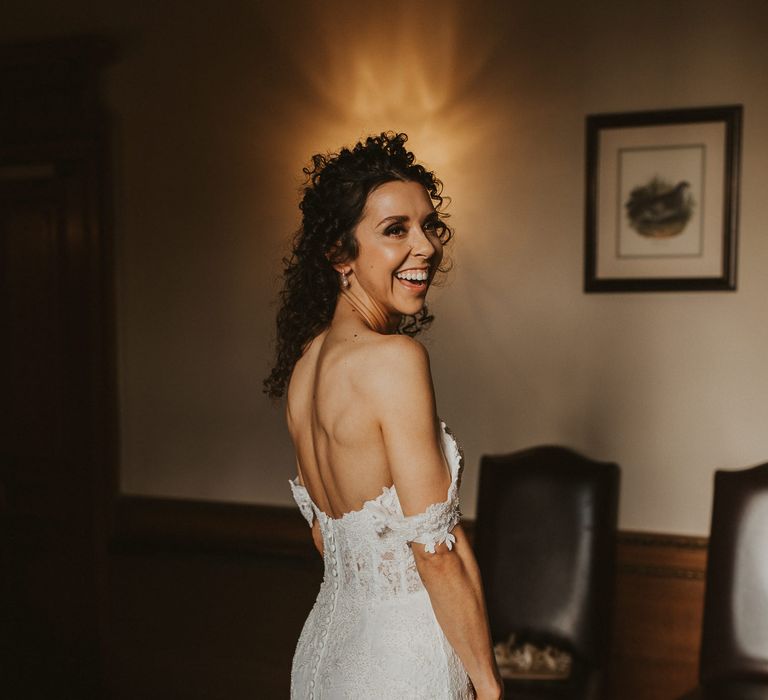 Bride with brown curly hair on her wedding morning in an off the shoulder lace wedding dress with buttons on the back 