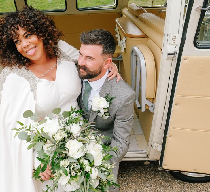 A Bride with curly hair sits on a grooms lap. She smiles and wears a wedding dress with a cape and holds a large white bouquet.