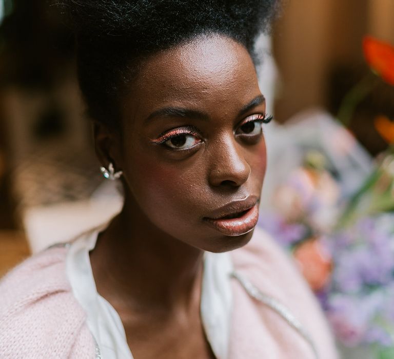 Wedding hairstyles for Black brides. A bride looks down to camera. She wears her afro hair swept back with neutral dewy make up and bright pink eyeliner.