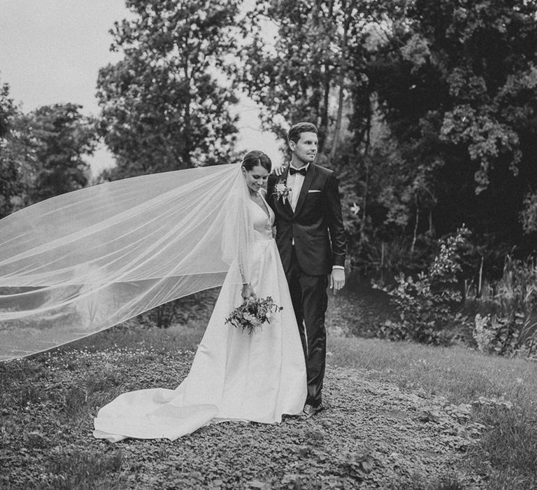 Bride in white Jesus Piero gown and cathedral length veil blowing in the wind stands with groom in black Tiger of Sweden tux in gardens at German wedding