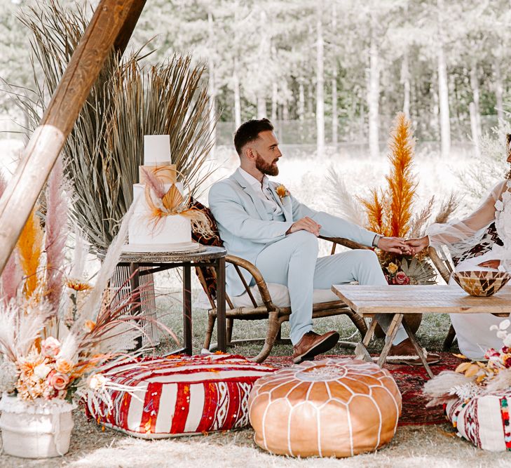 Bride & groom sit under tipi surrounded by brightly coloured cushions and wooden furniture