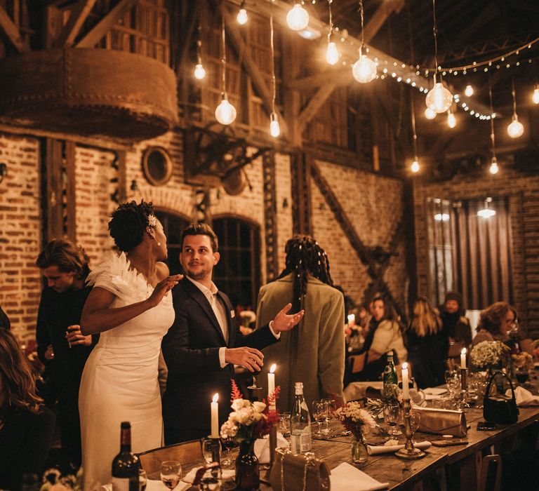 Bride in an off the shoulder wedding dress with appliqué shoulder detail taking to her guests at her rustic wedding reception 