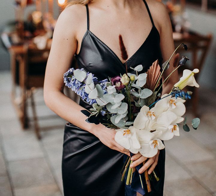 Bridesmaid in a leather thin strap dress holding an orchid and foliage wedding bouquet 