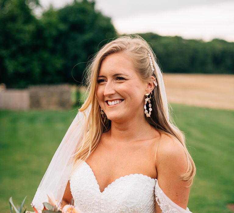 Bride smiling wearing off the shoulder lace bridal gown holding peach wedding bouquet with orchids, roses and King Proteas