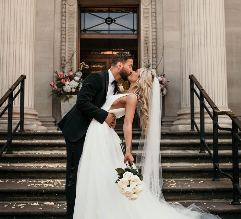 Groom leans back bride to kiss her on the steps of Marylebone Town Hall