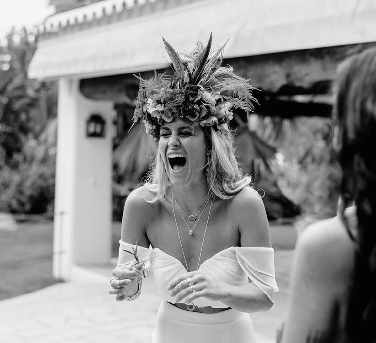 A bride in an oversized flower crown laughs. She wears a bardot neckline cropped top. Photography by Stephanie Shenton. 