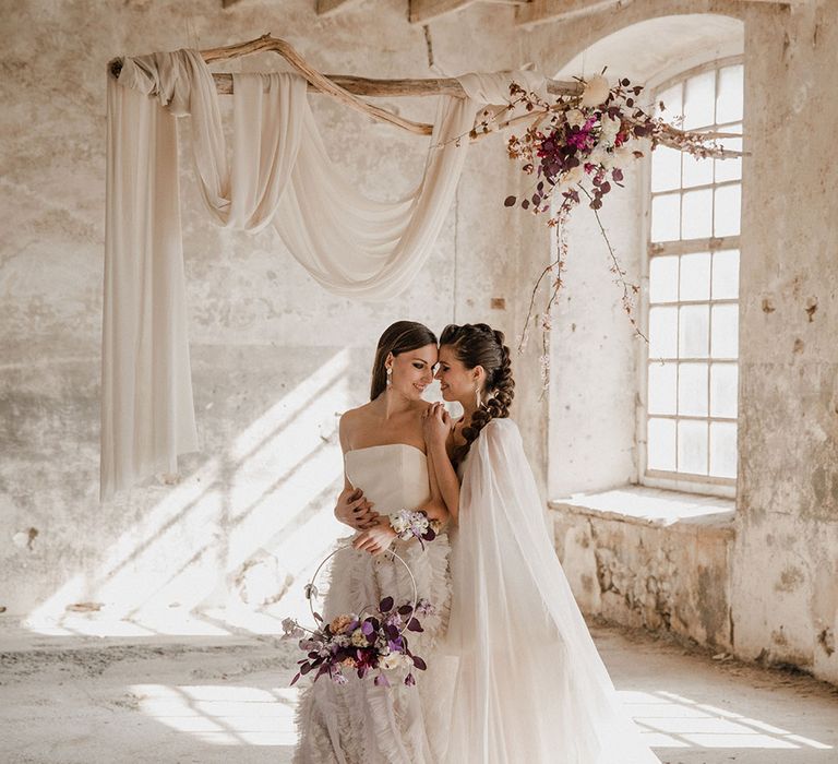 Bride with braided wedding hair wearing a wedding cape embracing her bride in a crop top and ruffle skirt holding a hoop bouquet under a drift wood handing display