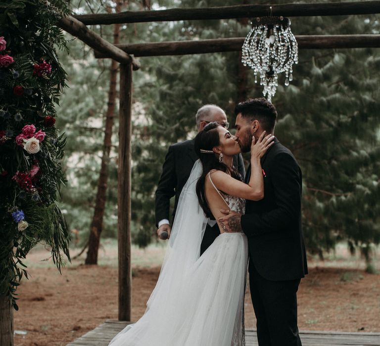 Bride and groom kiss as they get married, in moody romantic outdoor wedding ceremony with flowers and chandelier