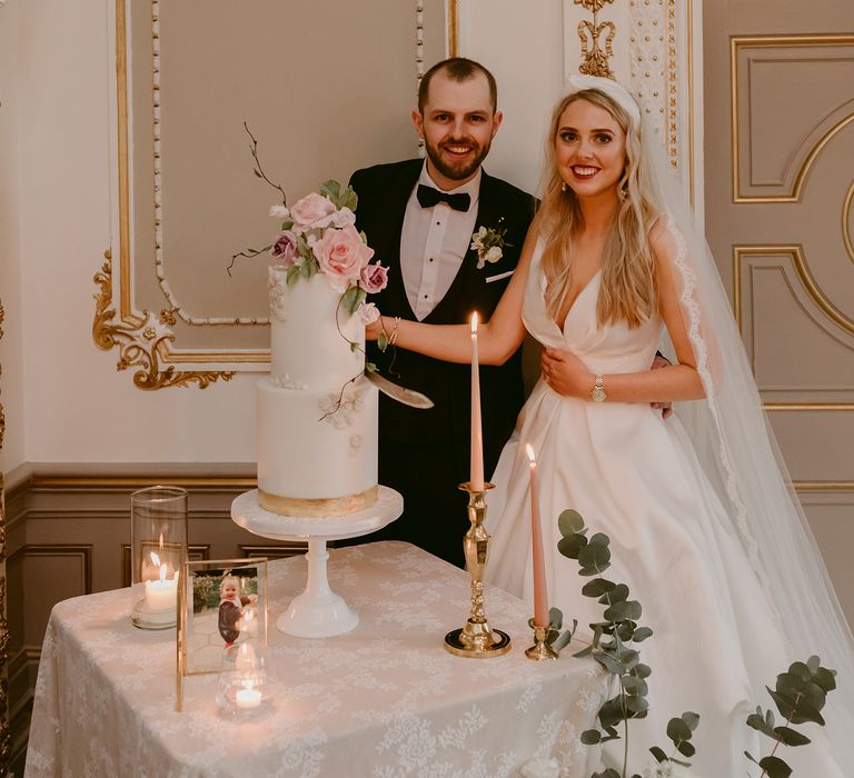 Bride & groom cut two tier wedding cake with white frosting and pink floral decor in white and gold gilded room