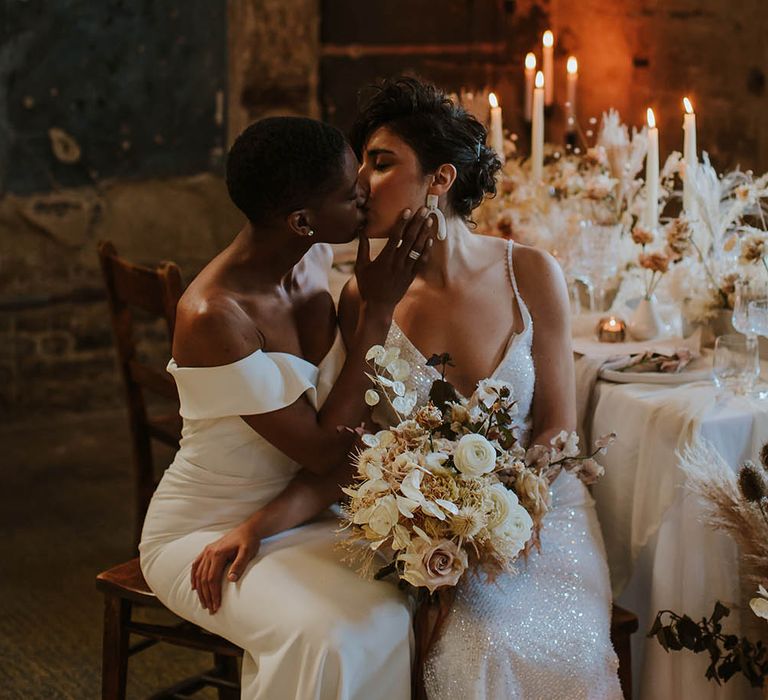 Bride in a Bardot fitted wedding dress kissing her bride in a sequin wedding dress with straps holding a dried and fresh flower bouquet 