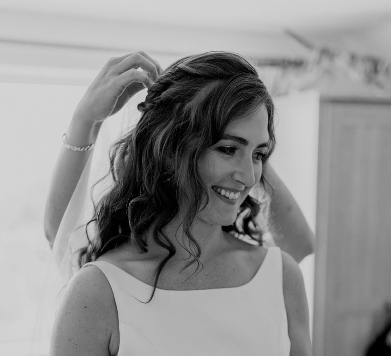 Smiling bride with long brown curled hair and sleeveless Elbeth Gillis wedding dress has her veil attached before home farm wedding