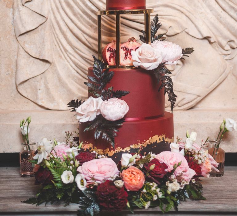 Luxury burgundy wedding cake with elevated third tier on a gold stand decorated with red, white and pink flowers, pomegranates and foliage.