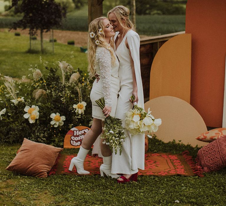 Bride in a jumpsuit with cape embracing her bride in a short wedding dress and bridal boots at an outdoor sixties wedding 