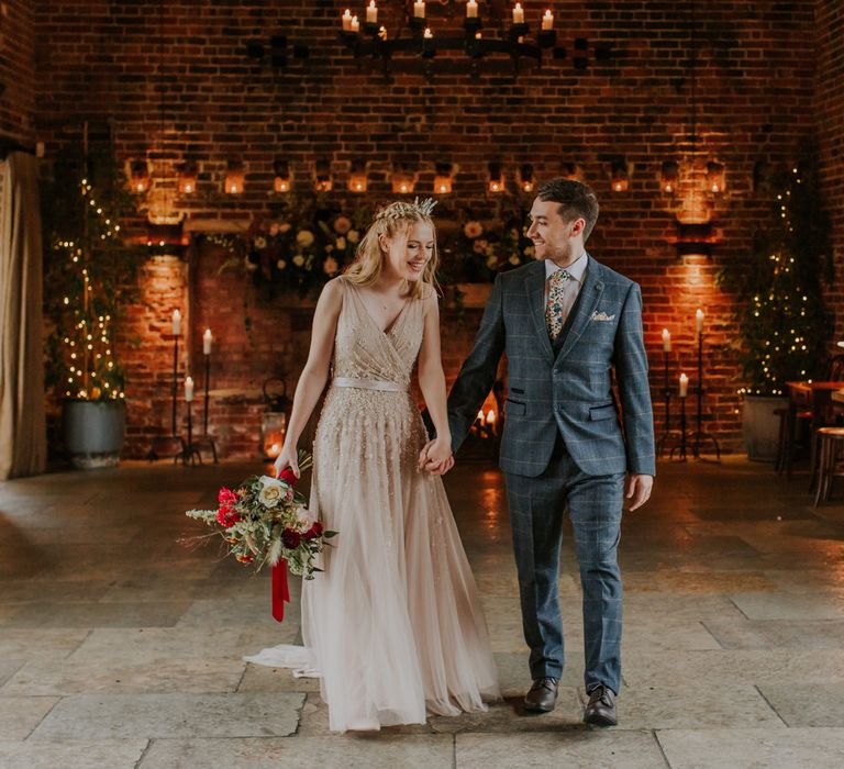 Bride in a blush coloured wedding dress and crown holding hands with her groom in a grey checked suit in the main reception room at Hazel Gap Barn