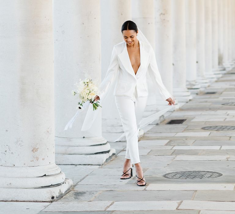 A bride at a black a white wedding wears a bespoke low cut white suit, a long veil and carries black and white wedding flowers.