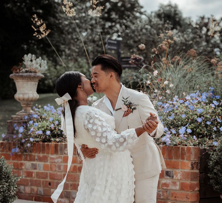 Groom in white shawl collared shirt and double breasted linen suit with brown shoes and floral buttonhole dances with bride in white long mesh sleeve spotted Vagabond wedding dress and bridal hair bow outside in gardens at Wasing Park wedding