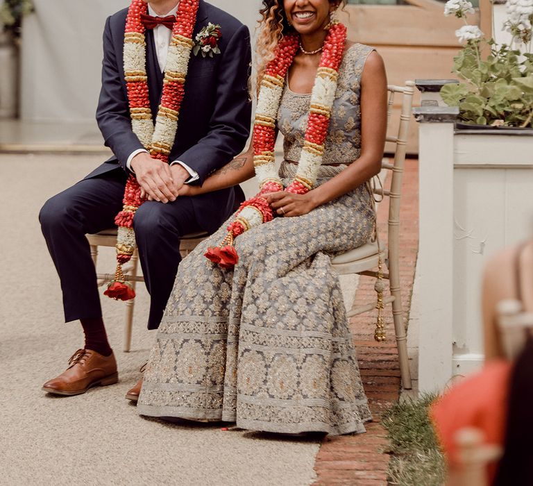 Bride & groom sit during wedding ceremony with colourful garlands around their necks | Joshua Gooding Photography