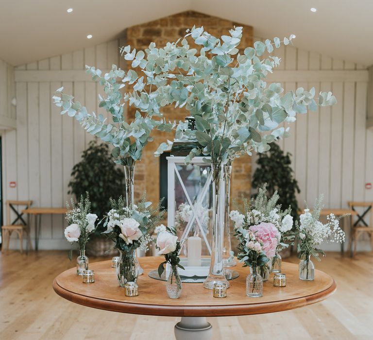 Round wooden table with florals in glass bottles, large dried leaf stems and glass candle holder for summer wedding at Primrose Hill Farm