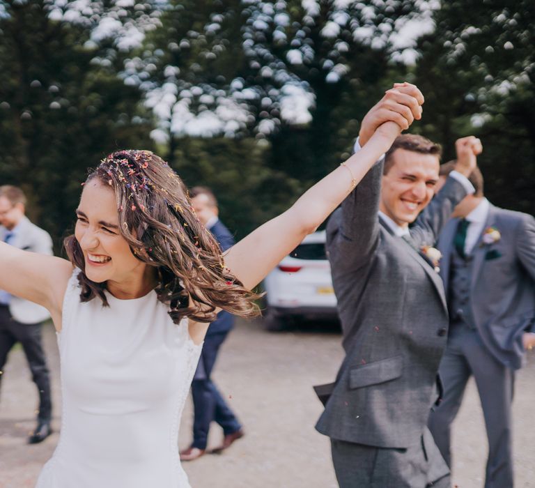 White, brunette bride in white dress with arms in the air holds hands with white groom in grey suit during confetti exit