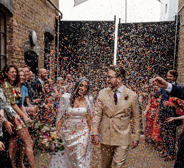 Confetti moment at the Loft Studios London with bride in a fitted wedding dress and feather cape and groom in a beige suit with double breasted jacket 