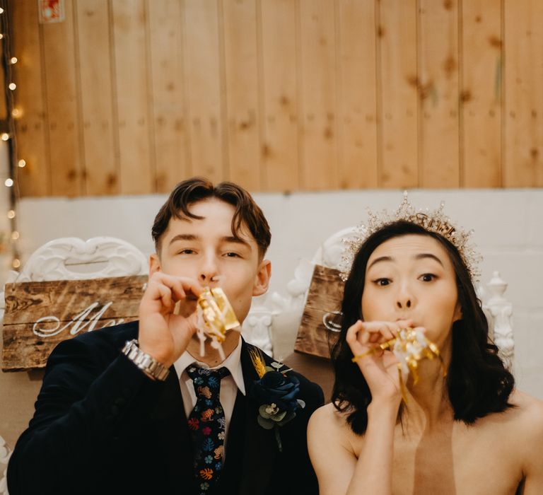 Groom in blue suit and floral tie and bride in strapless wedding dress and headpiece blow party blowers during rustic wedding ceremony in Lancashire