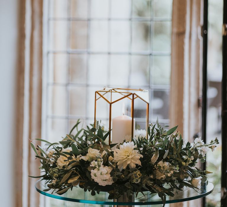 Candle in glass candle holder surrounded by wreath of white and green flowers on glass table at Notley Abbey wedding