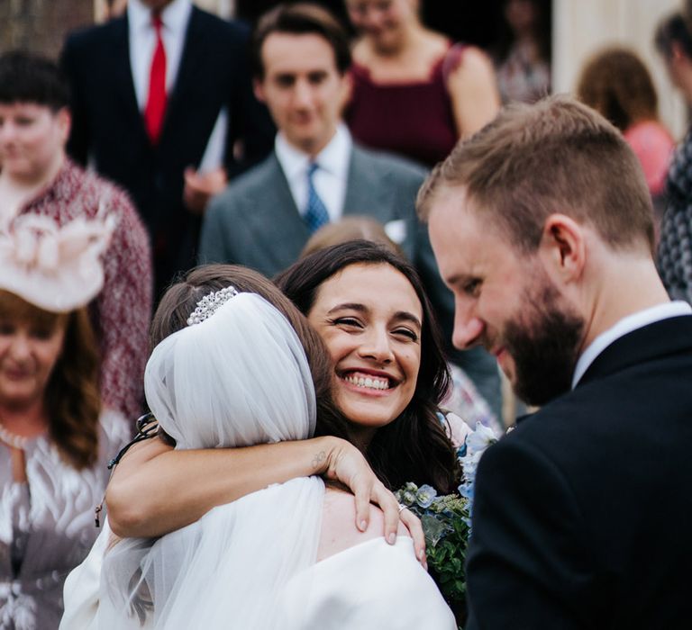 Bride hugs wedding guest as groom smiles on their wedding day