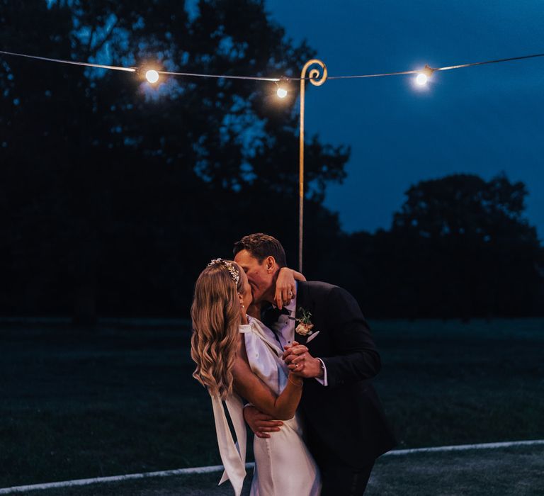 Bride in satin halterneck wedding dress with train and floral headband hugs groom in black tie as they dance outside by hanging bulb lights