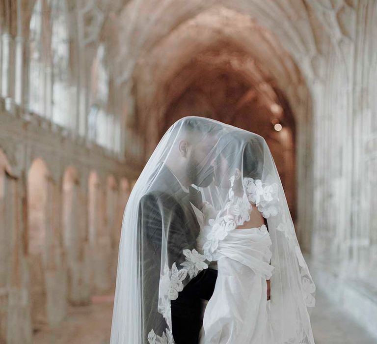 Bride and groom kissing in a medieval wedding venue corridor with a lace edge veil over them 