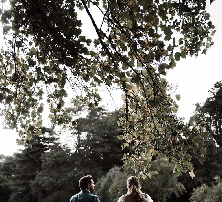 Groom in blue suit walks holding hands with bride in low back embroidered Victoria Sanders wedding dress with long sleeves through the grounds of Iscoyd Park