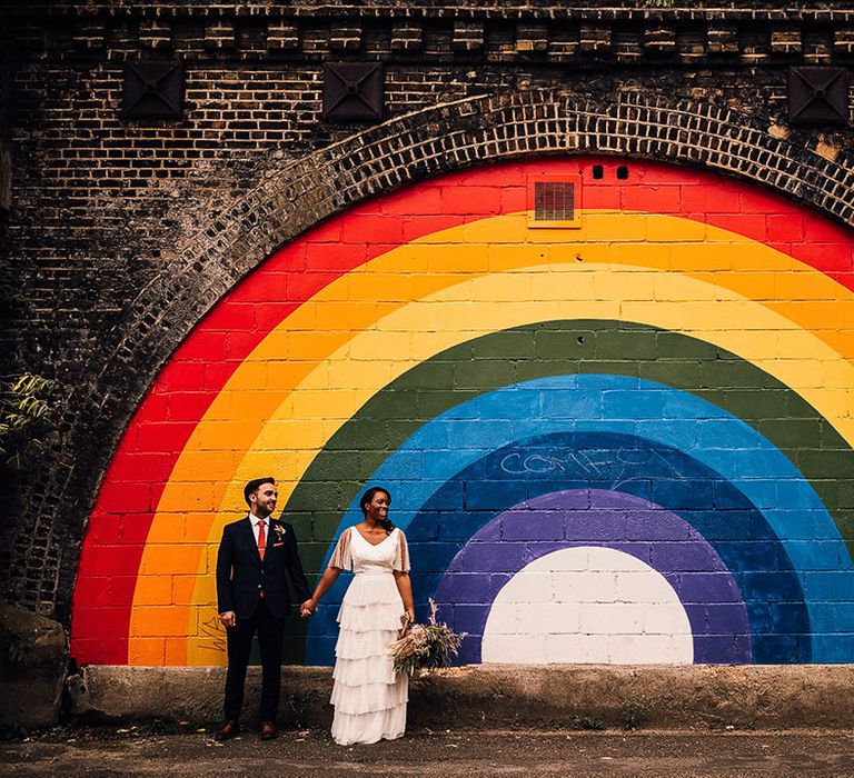 Black bride in Story Of My Dress bridal separates and groom in a navy suit standing in front of a giant street art rainbow motif in Brixton 