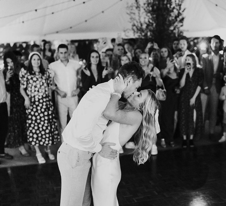 Bride in low back satin dress kisses groom in white shirt and suit trousers on the dance floor at marquee wedding reception