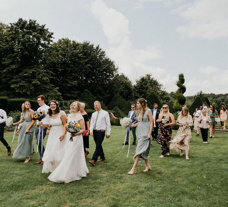 Blonde and brunette brides laugh together as their wedding party walks with them around the grounds of the barn wedding venue