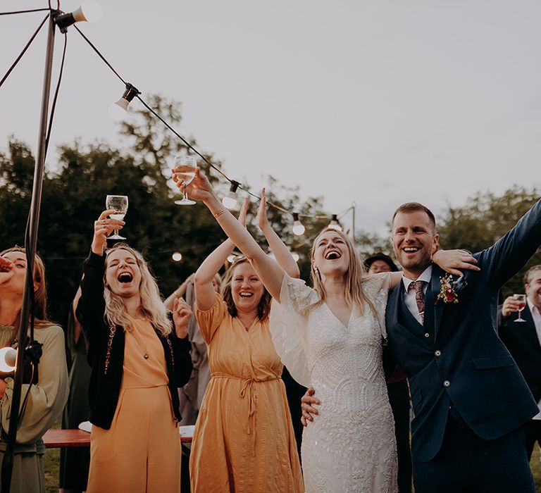 Bride in beaded wedding dress and groom in blue three piece suit put their hands in the air at reception