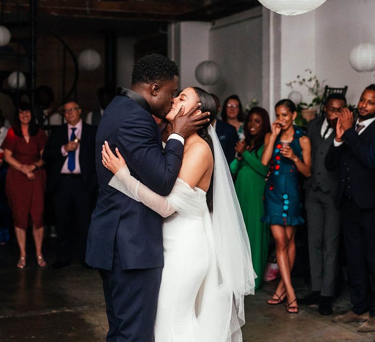 Bride and groom kiss while having their first dance as wedding guests clap and watch adoringly