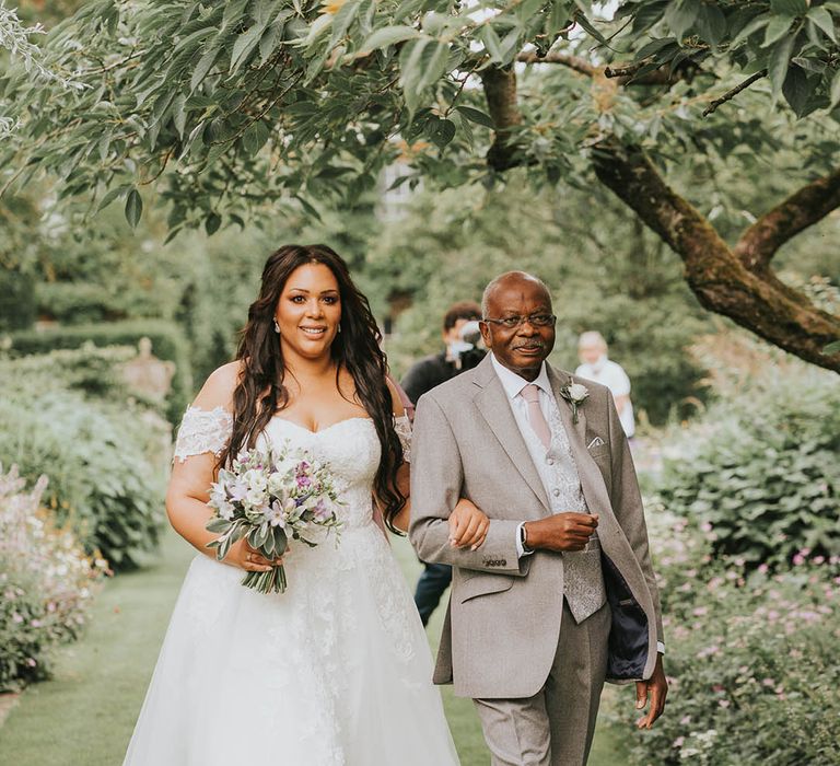 Father of the bride in light grey suit, patterned waistcoat and pink tie walks the bride down the aisle in lace off the shoulder wedding dress